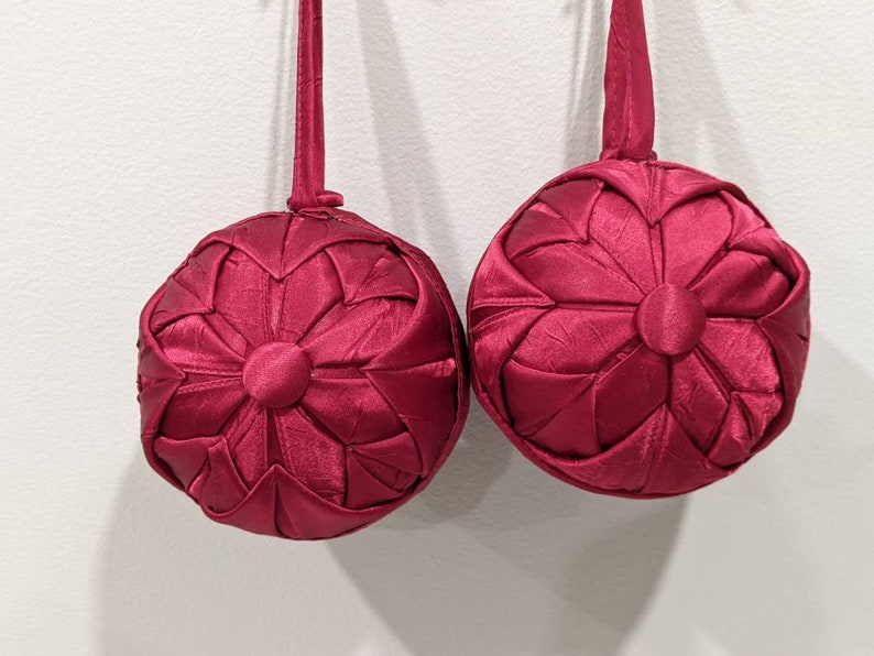May include: Two burgundy fabric Christmas ornaments with a starburst design. The ornaments are hanging from a white wall.