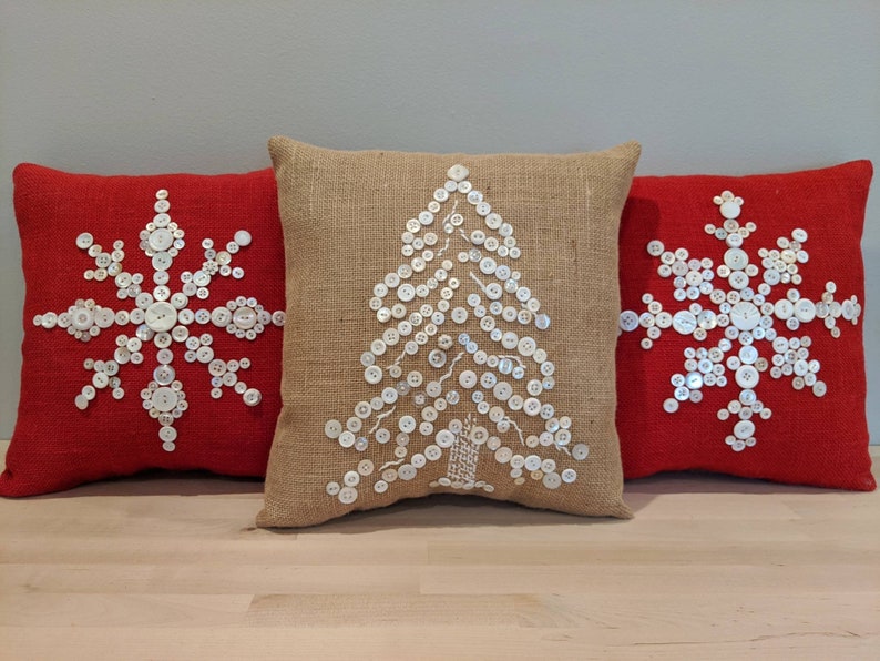 May include: Three decorative throw pillows with a Christmas theme. Two pillows are red with white button snowflakes and one pillow is burlap with a white button Christmas tree.