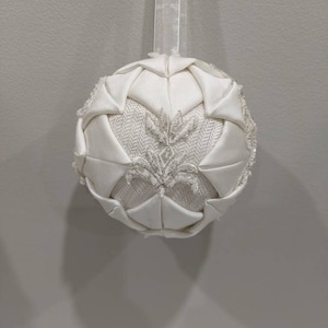 May include: A white, fabric Christmas ornament with a beaded design. The ornament is shaped like a sphere and has a loop at the top for hanging.