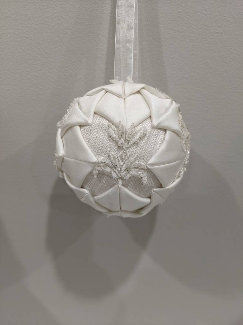 May include: A white, fabric Christmas ornament with a beaded design. The ornament is shaped like a sphere and has a loop at the top for hanging.