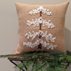 May include: A burlap pillow with a white button tree design. The pillow is decorated with white buttons of various sizes, arranged in the shape of a tree.