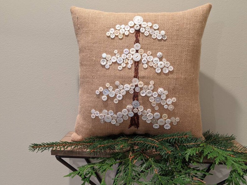 May include: A burlap pillow with a white button tree design. The pillow is decorated with white buttons of various sizes, arranged in the shape of a tree.
