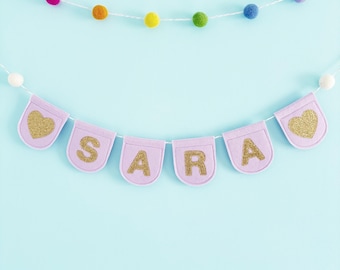 May include: A pink felt banner with the name "SARA" spelled out in gold glitter letters. The banner is strung with white thread and decorated with colorful pom-poms.