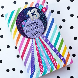 May include: Purple and blue fabric birthday badge with a silver glitter number 13, stars, and the text "Happy Birthday Bella". The badge is attached to rainbow striped wrapping paper with white polka dots.