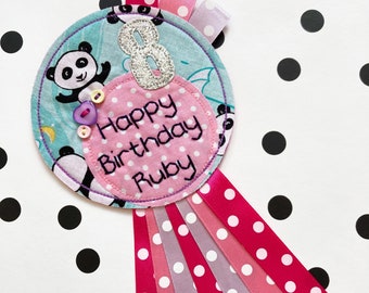 May include: A pink and blue fabric birthday badge with a panda design, a silver glitter number 8, and the text "Happy Birthday Ruby". The badge is attached to a set of pink and white polka dot ribbons.