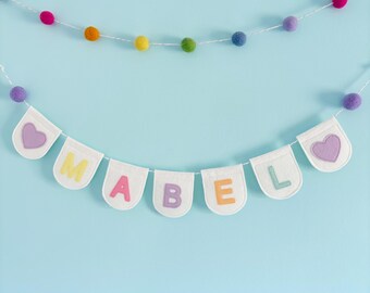 May include: A white felt banner with the name "MABEL" spelled out in colorful felt letters. The banner is strung with a white string and decorated with a string of colorful pom-poms.