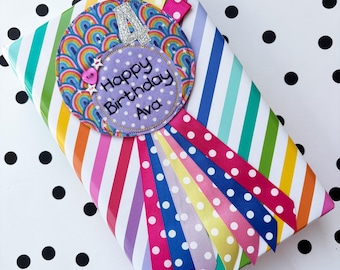 May include: A purple and rainbow fabric circle with the text "Happy Birthday Ava" and the number "4" in silver glitter. The circle is attached to a colorful ribbon with white polka dots.