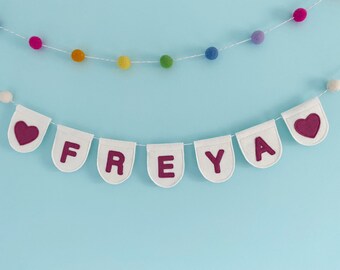 May include: White felt banner with the name "FREYA" in pink felt letters and two pink felt hearts. The banner is strung with a white string and white pom-poms.