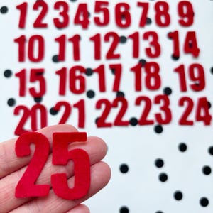 May include: Red felt numbers 1 through 25 on a white background with black polka dots. The number 25 is being held in a hand.