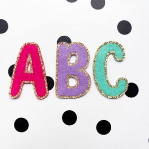 May include: Three felt letters, "ABC", in bright colors: pink, purple, and teal. Each letter has a gold glitter outline. The background is white with black polka dots. These letters are suitable for crafts and educational projects.