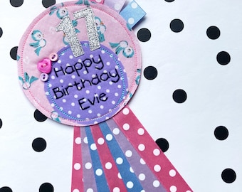May include: A pink and purple fabric birthday badge with the number 17, the words "Happy Birthday Evie" and a pink heart button. The badge is attached to a ribbon with pink, blue, and purple polka dots.