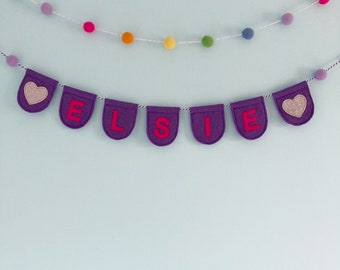 May include: Purple felt banner with the name "ELSIE" in pink letters and two silver glitter hearts. The banner is strung with white thread and hangs against a light blue wall.