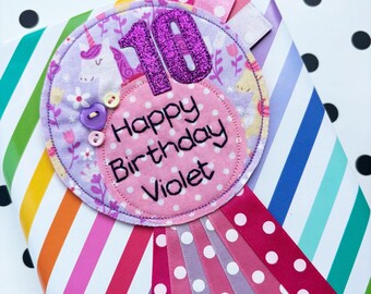 May include: A pink and purple fabric birthday badge with a unicorn design and the number 10 in glitter. The badge says "Happy Birthday Violet" and is attached to a fan of pink and white polka dot ribbons.