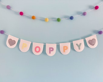 May include: A white felt banner with the name "POPPY" spelled out in rainbow colors. The banner is strung with a white string and has small, colorful pom-poms on either side.