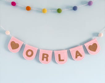 May include: Pink felt banner with the name "ORLA" in gold glitter letters and two gold glitter hearts on either end. The banner is strung with white thread and is hanging on a light blue wall.
