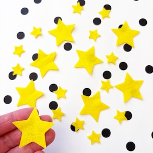 May include: A collection of yellow felt star shapes of different sizes, alongside black polka dots, on a white background. One star is held in a hand. The stars are a vibrant yellow, and the dots are black. The image is a top-down view.