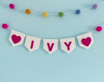 May include: White felt banner with the name "IVY" in pink felt letters and two pink felt hearts on either end. The banner is strung with a white string with colorful pom-poms.