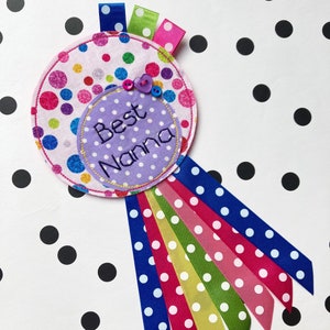 May include: A handmade fabric rosette with the words "Best Nanna" embroidered on a purple polka dot circle. The rosette features colorful polka dot fabric, ribbons, and decorative buttons. Ribbons in blue, pink, green, and red extend from the rosette.