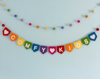 May include: A rainbow felt banner with the words "COMFY KIDS" and hearts. The banner is strung on a white string with colorful pom-poms.