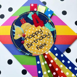 May include: A colorful birthday badge with a dinosaur, presents, and the number 4. The badge says "Happy Birthday Felix" in yellow embroidery on a yellow circle with white polka dots.