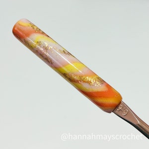 May include: A close-up of a crochet hook with a swirl pattern in orange, yellow, white, and gold glitter. The hook is labeled "5.0".