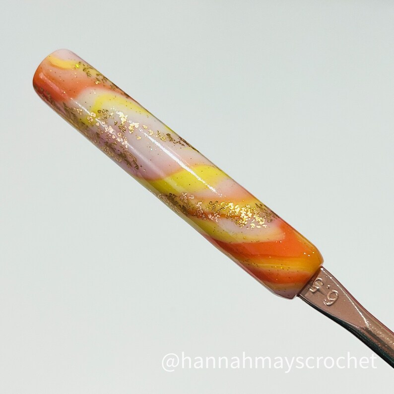 May include: A close-up of a crochet hook with a swirl pattern in orange, yellow, white, and gold glitter. The hook is labeled "5.0".