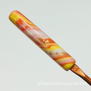 May include: A close-up of a crochet hook with a marbled orange, yellow, and white handle. The handle is decorated with gold flakes. The hook is made of metal and has a copper finish. The text "@hannahmayscrochet" is visible at the bottom of the image.