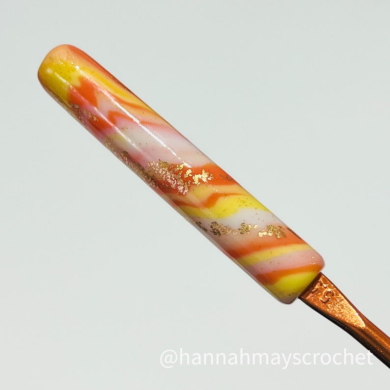 May include: A close-up of a crochet hook with a marbled orange, yellow, and white handle. The handle is decorated with gold flakes. The hook is made of metal and has a copper finish. The text "@hannahmayscrochet" is visible at the bottom of the image.