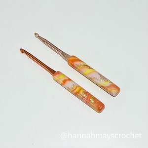 May include: Two crochet hooks with orange, yellow, and white marbled handles. The hooks are made of metal and have a shiny finish.