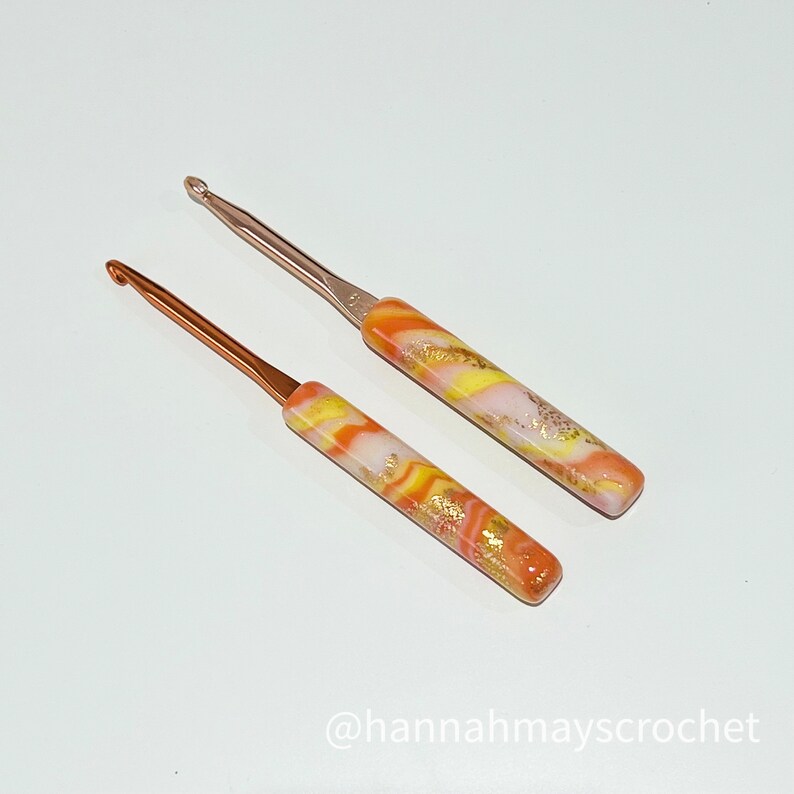 May include: Two crochet hooks with orange, yellow, and white marbled handles. The hooks are made of metal and have a shiny finish.