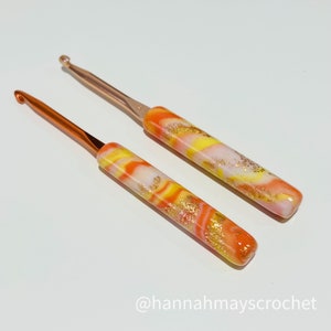 May include: Two colorful crochet hooks with orange, yellow, and white marbled handles and gold flecks. The hooks have a rose gold metal tip.