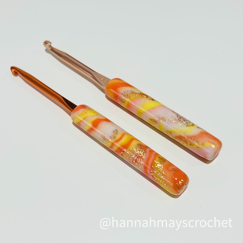 May include: Two colorful crochet hooks with orange, yellow, and white marbled handles and gold flecks. The hooks have a rose gold metal tip.