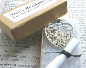 May include: A silver heart-shaped pendant necklace with a small, round, yellow stone in the centre. The necklace is in a small, white box with a black ribbon. The box is sitting on a page of a book with text about Jesus.