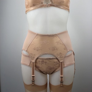 May include: A vintage-style lingerie set in a soft peach color, featuring a bra and high-waisted garter belt. The bra has a delicate floral lace design on the cups, with adjustable straps. The garter belt has matching lace details and suspender straps.