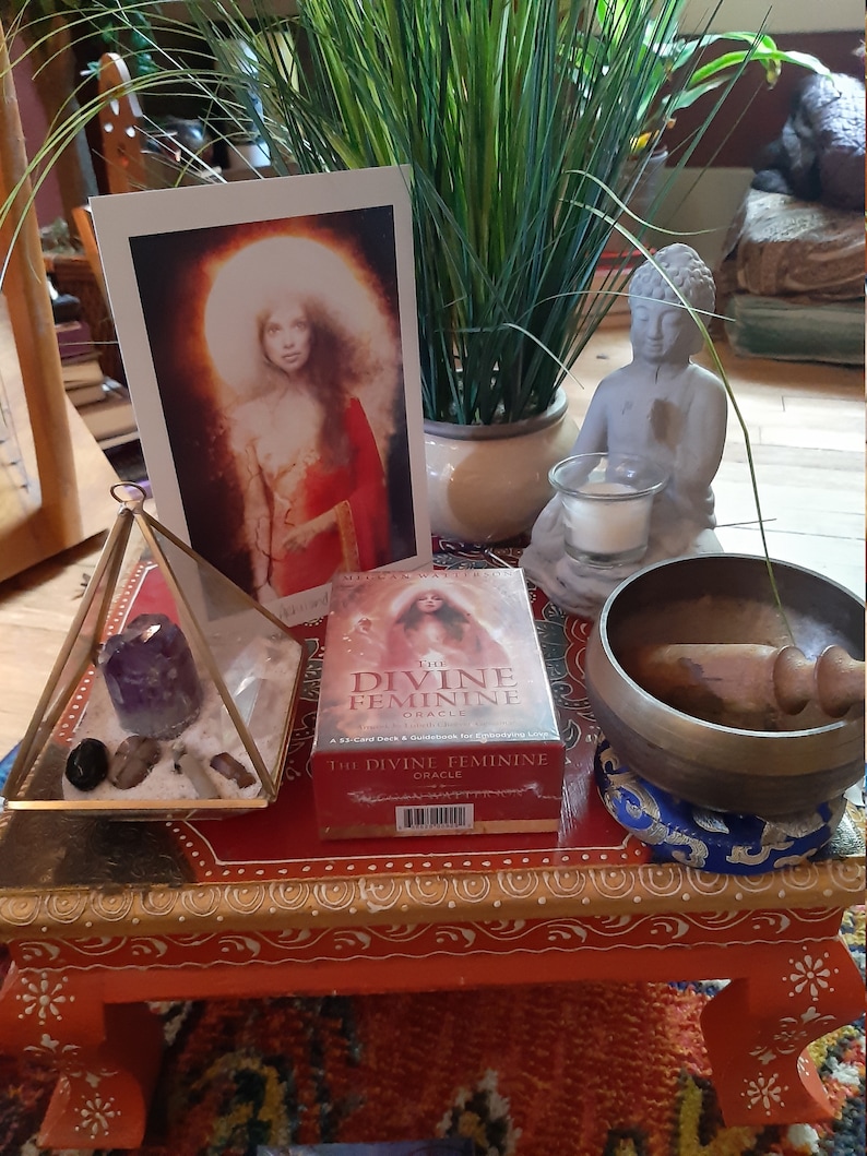 K&ouml;nnte beinhalten: Ein roter Holztisch mit goldenen Details, auf dem eine Kristallpyramide mit Amethystkristallen, ein Orakelkartenspiel mit dem Titel "The Divine Feminine Oracle", eine kleine wei&szlig;e Buddha-Statue, eine Kerze in einem Glasgef&auml;&szlig; und eine Klangschale mit einem Holzschl&auml;gel stehen.