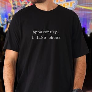 Op de afbeelding: Zwart t-shirt met de witte tekst "apparently, i like cheer". Het shirt is gemaakt van een zacht materiaal en heeft korte mouwen. De tekst is in een eenvoudige, schreefloze lettertype.