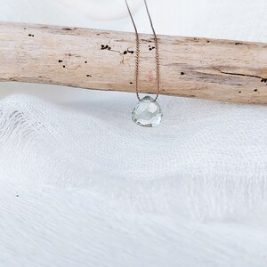 May include: A delicate necklace with a light green gemstone pendant. The pendant is faceted and has a teardrop shape. The necklace is made of thin, brown cord.