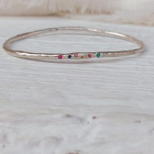 Op de afbeelding: Een zilveren armband met een getextureerd oppervlak. De armband is versierd met kleine, kleurrijke edelstenen in rood, blauw, roze, oranje en groen. De armband wordt gepresenteerd op een wit houten oppervlak.
