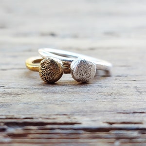 Op de afbeelding: Twee eenvoudige zilveren en gouden ringen met getextureerde ronde toppen. De ringen zijn op elkaar gestapeld op een houten oppervlak.