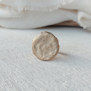 May include: A gold-colored ring with a textured, organic design. The ring has a rounded, irregular shape, and the surface appears to have a matte finish. The ring is set against a white background with a soft, neutral fabric in the background.