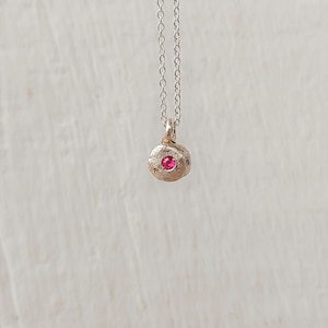 Könnte beinhalten: Eine silberne Halskette mit einem kleinen, strukturierten Anhänger. Der Anhänger hat einen hellrosa Edelstein in der Mitte. Die Kette ist zart und hängt vertikal vor einem neutralen Hintergrund. Der Schmuck ist schlicht und elegant.