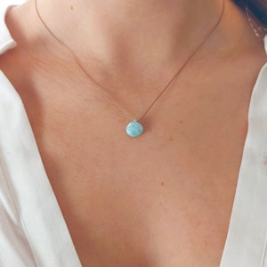 May include: A delicate necklace featuring a small, teardrop-shaped light blue stone pendant with white marbling, suspended from a thin, metallic rose gold-colored chain that rests against the collarbone area of a white collared shirt.