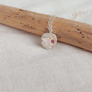 Peut inclure: Un collier en argent avec un pendentif texturé orné d'une petite pierre précieuse rose. Le collier est présenté sur un morceau de bois flotté.