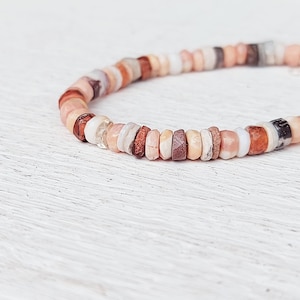 Puede incluir: Una pulsera de cuentas planas y redondas en tonos tierra. Las cuentas son de diferentes colores: rosa, marrón, blanco y beige. La pulsera está hecha de pequeñas cuentas planas y redondas, probablemente ensartadas en un cordón elástico.