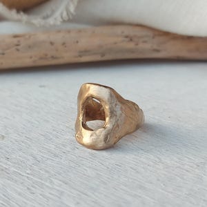 Könnte beinhalten: Ein goldfarbener Ring mit matter, texturierter Oberfläche und unregelmäßiger, organischer Form. Das Band weist eine markante, offene, hohle Mitte auf, die einen deutlichen Hohlraum innerhalb der Metallstruktur erzeugt und ein raues Aussehen zeigt.