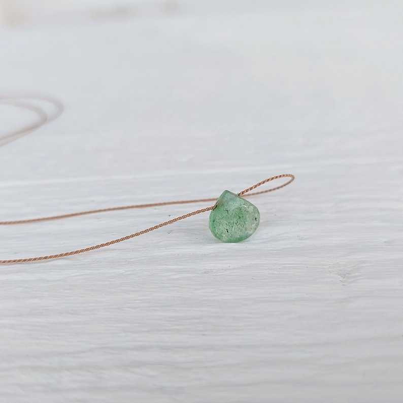 May include: A green gemstone pendant on a brown cord necklace. The pendant is a teardrop shape and has a rough, natural texture.