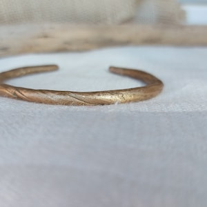 Op de afbeelding: Een bronzen manchetarmband met een getextureerd, gedraaid ontwerp. De open armband wordt getoond op een witte ondergrond, waarbij de metallic glans wordt benadrukt. Dit sieraad is een eenvoudig maar elegant accessoire.
