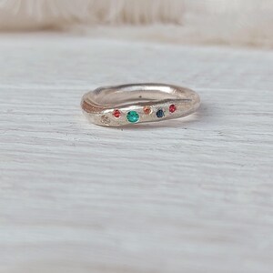 Può includere: Un anello d'argento con una superficie strutturata. L'anello è adornato da una serie di piccole pietre preziose colorate, tra cui rosso, verde, arancione e blu. L'anello è posto su una superficie di legno chiaro.