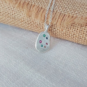 May include: A silver necklace with a small, oval-shaped pendant. The pendant is set with a variety of colorful gemstones, including red, green, and blue.