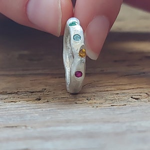 May include: A silver ring with a textured surface, adorned with five colorful gemstones. The gemstones are green, light blue, yellow, and red. The ring is held between two fingers.
